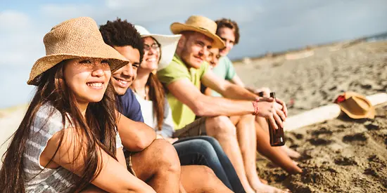 Gruppen-Reiseversicherungen - Eine Gruppe junger Menschen sitzen am Strand. Die Sonne schneit.
