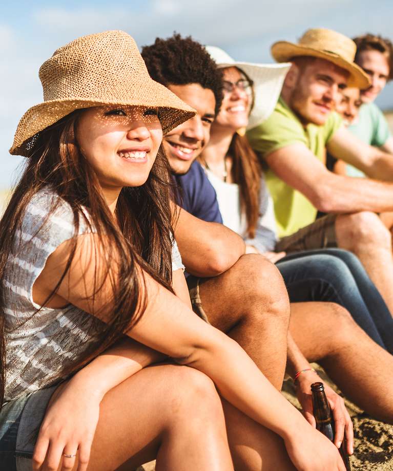 Gruppen-Reiseversicherungen - Eine Gruppe junger Menschen sitzen am Strand. Die Sonne schneit.
