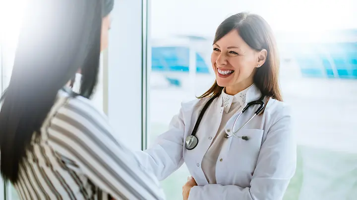 Eine Frau in einer gestreiften Bluse steht vor einer Ärztin. Die Ärztin lächelt die Frau an. 