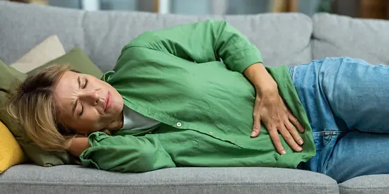 Eine Frau mit grüner Bluse liegt auf dem Sofa und hält sich den Bauch vor Schmerzen.