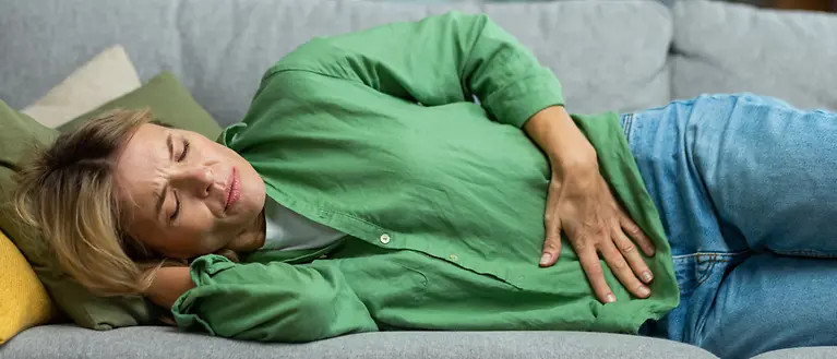 Eine Frau mit grüner Bluse liegt auf dem Sofa und hält sich den Bauch vor Schmerzen.