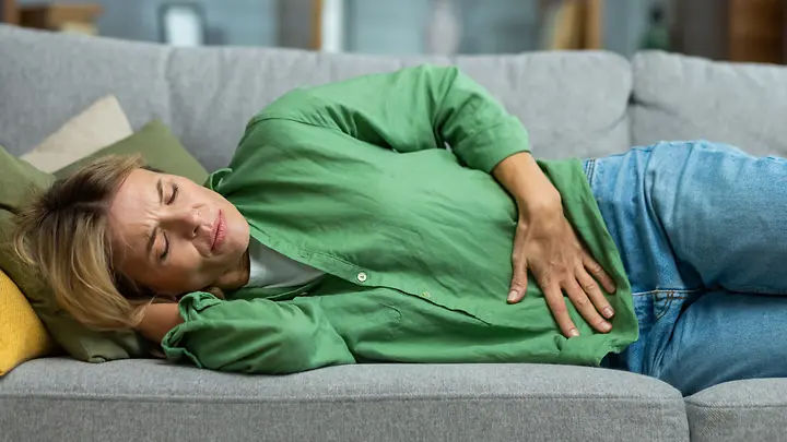 Eine Frau mit grüner Bluse liegt auf dem Sofa und hält sich den Bauch vor Schmerzen.